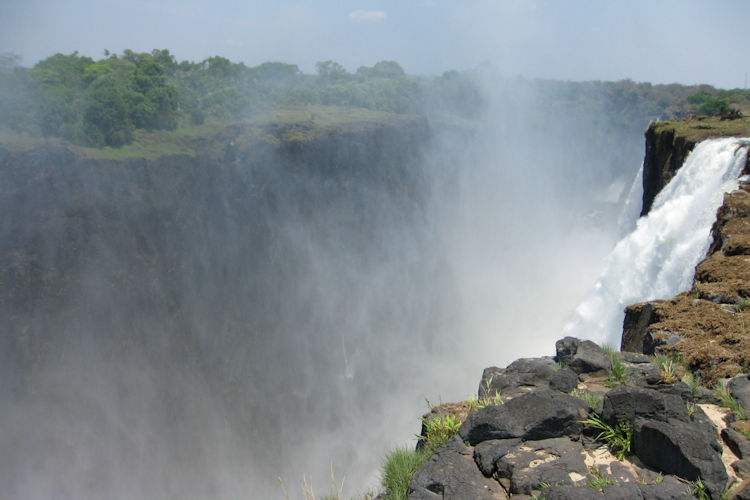 An aerial view of Victoria Falls showcases the magnificent waterfalls cascading into the Batoka Gorge, with mist rising dramatically above the landscape. Surrounding the falls are lush green areas, highlighting the natural habitat of wildlife and the breathtaking scenery that attracts visitors to this popular tourist attraction in Livingstone.