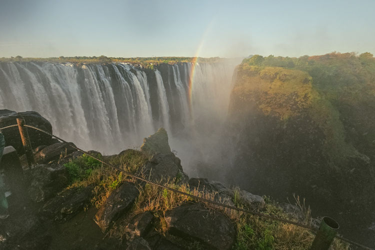 A breathtaking panoramic view of Victoria Falls showcases the mighty Zambezi River cascading down, with mist rising dramatically into the sky, creating a vibrant rainbow in the spray. This iconic natural wonder, located within Victoria Falls National Park, is a must-see attraction for visitors exploring the beauty of Southern Africa.