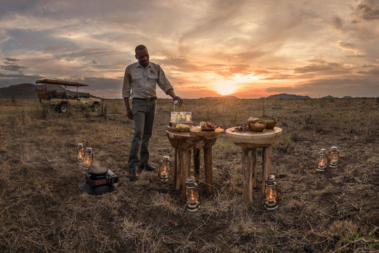 An aerial view captures the stunning African savanna landscape at golden hour, featuring scattered acacia trees and distant mountains, reminiscent of the serene beauty found in the Madikwe Game Reserve. This picturesque scene embodies the essence of a luxury safari experience, inviting visitors to explore the wild and unwind in intimate luxury lodges.