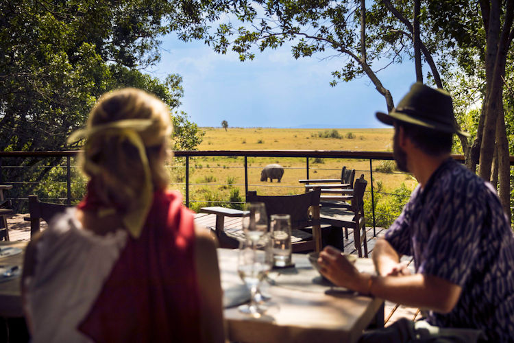 A romantic couple sits on a private deck, sipping drinks as they watch the vibrant sunset over the African savanna, adorned with blooming wildflowers. The scene captures the stunning backdrop of Amboseli National Park, creating an unforgettable moment perfect for a romantic safari getaway.