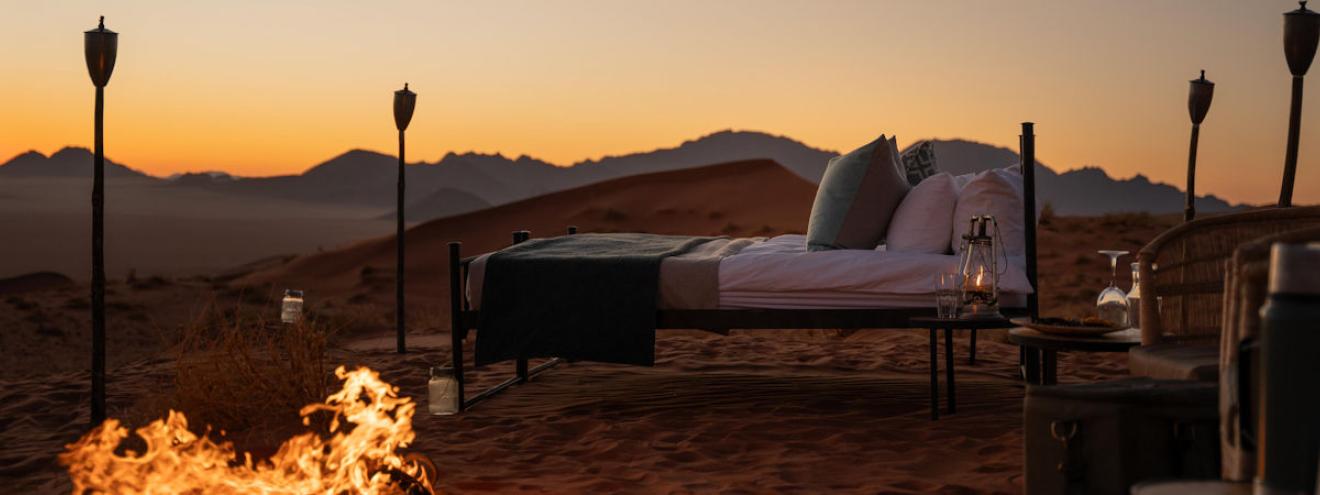 A luxuriously dressed star bed set on a raised timber deck amid the rust-red dunes of the Namib Desert at sunset, flanked by tall tiki torches and a glowing campfire in the foreground, with the silhouette of desert mountains bathed in deep amber and golden light stretching across the horizon behind.