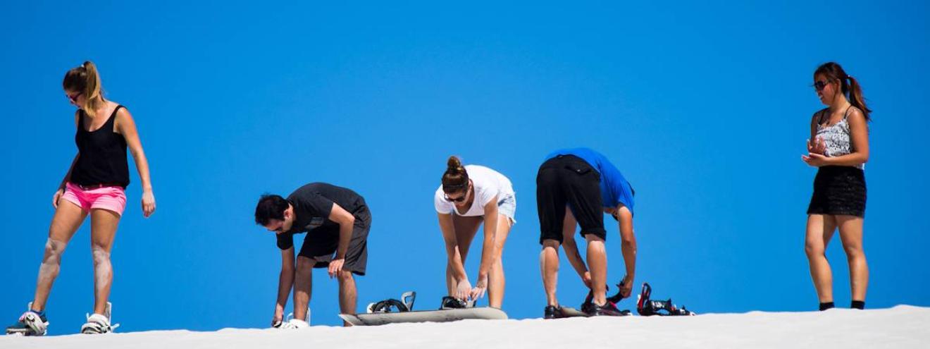 A group of tourists strapping on sandboards at the top of white sand dunes on a South Africa day tour near Cape Town
